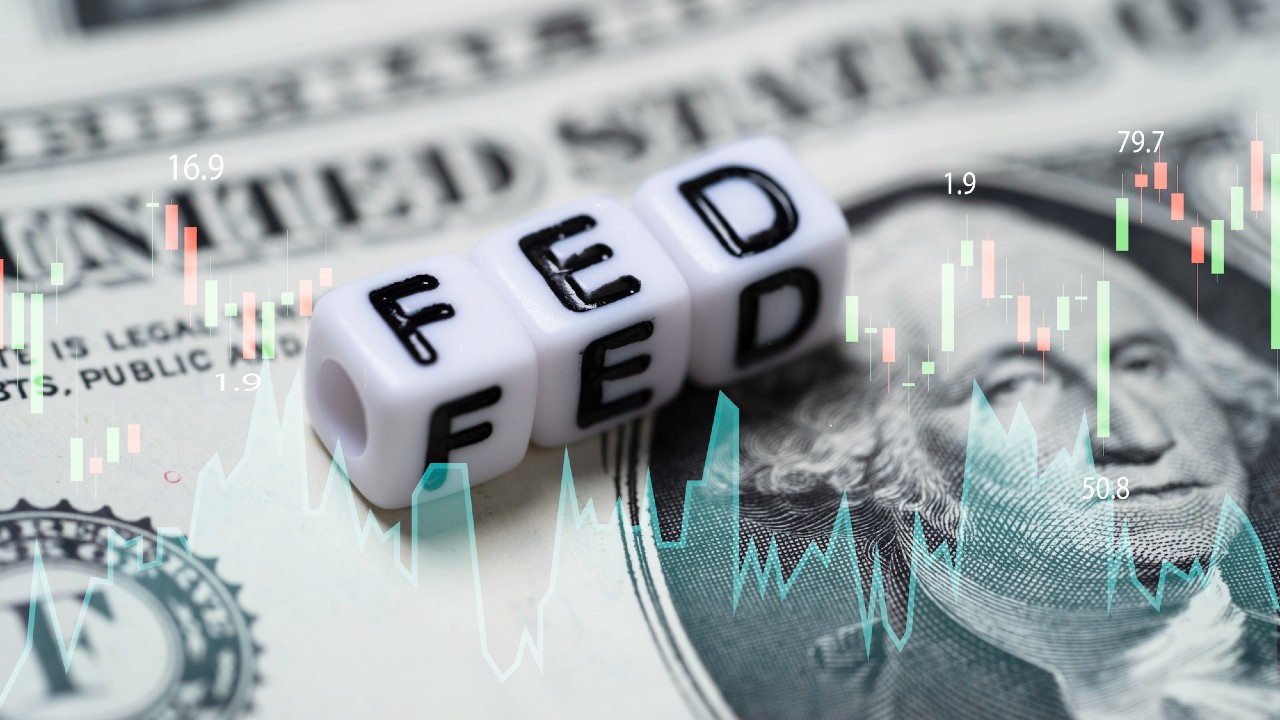 Cubi con le lettere che compongono la parola "FED" sono appoggiati su una banconota da un dollaro statunitense. Un grafico con linee e numeri sovrappone l'immagine, suggerendo un'analisi finanziaria o economica.