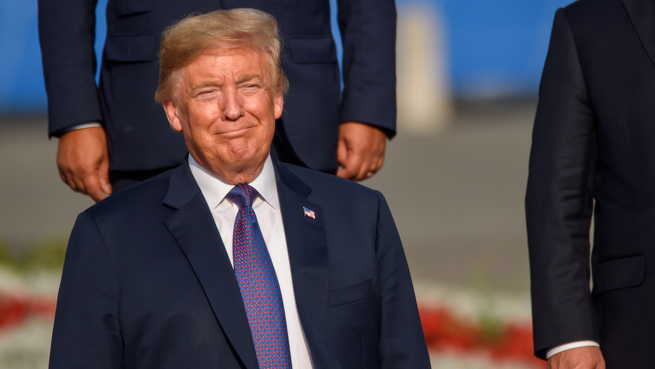 Un uomo parla su un podio con un cartello "TRUMP" e uno striscione "Make America Great Again!". Gesticola con la mano verso la testa. Una folla di persone è seduta sullo sfondo.