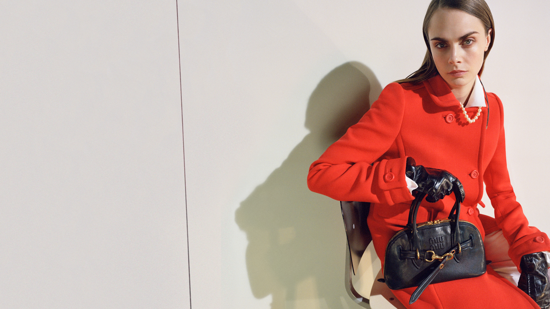 Il lusso fa i conti con Prada e Miu Miu (che cresce del 100%) Una persona che indossa un cappotto rosso acceso e guanti di pelle nera è seduta su una sedia contro un muro semplice. Tiene una borsa nera alla moda sulle ginocchia e ha i capelli lisci e divisi. La scena è minimalista e alla moda.