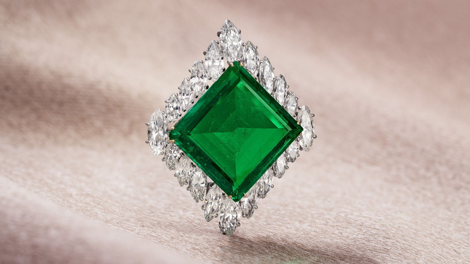 Un grande smeraldo dal taglio quadrato è circondato da una disposizione geometrica di diamanti scintillanti, creando un gioiello intricato e lussuoso su uno sfondo morbido e strutturato.
