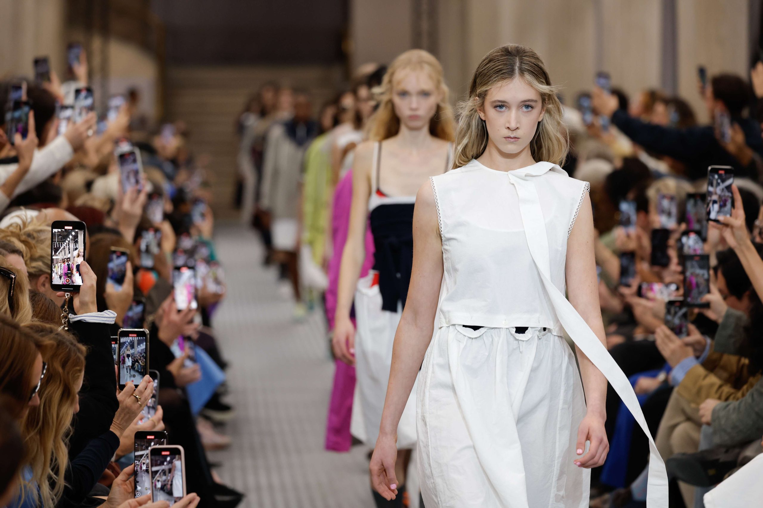 Miu Miu al top della classifica Lyst Le modelle sfilano su una passerella di moda indossando abiti eleganti. Molti membri del pubblico da entrambe le parti tengono in mano i telefoni per immortalare l'evento. L'ambiente è luminoso, con toni neutri sullo sfondo.