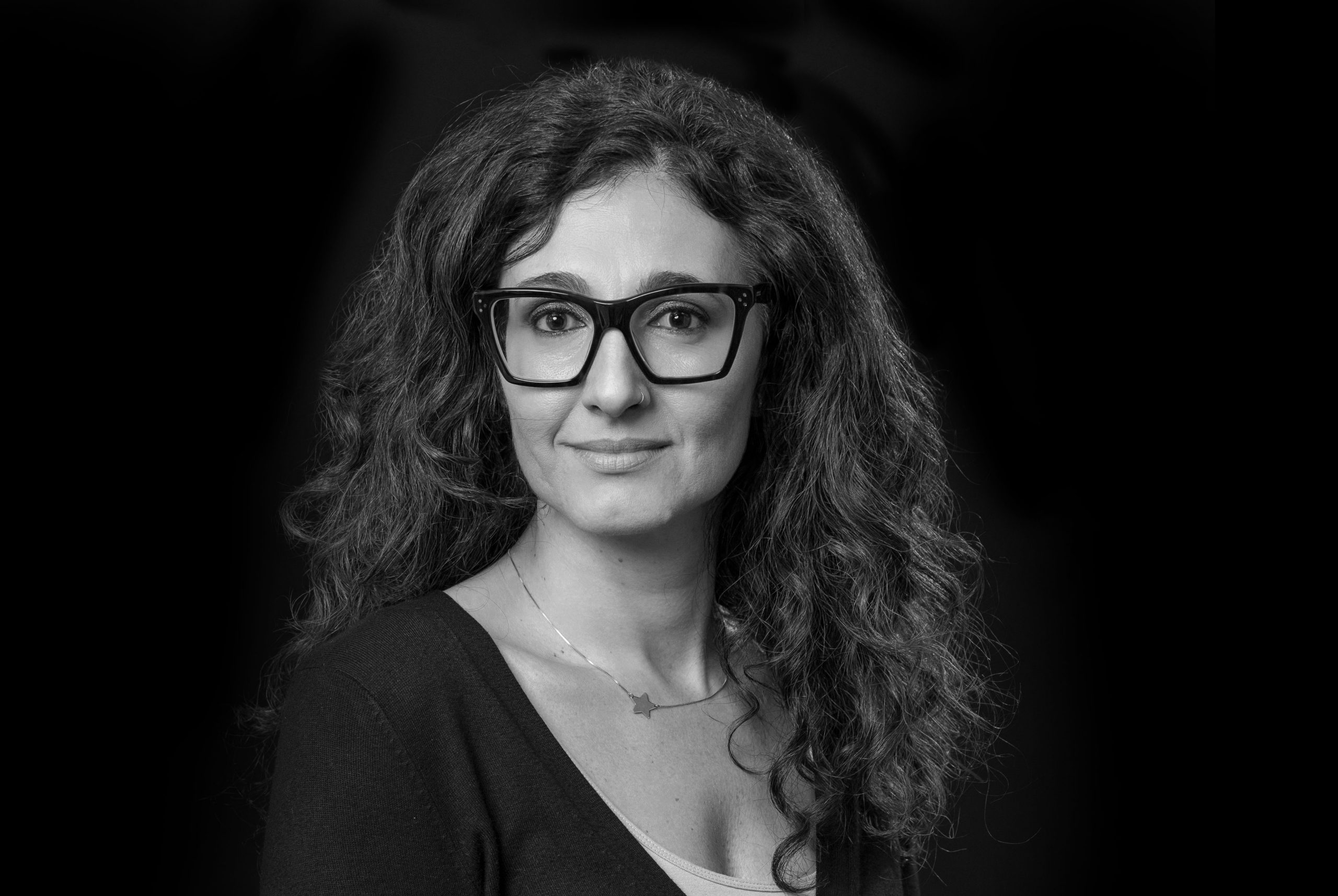 Ritratto in bianco e nero di una persona con lunghi capelli ricci e grandi occhiali, che indossa una collana e un top scuro, su uno sfondo scuro.