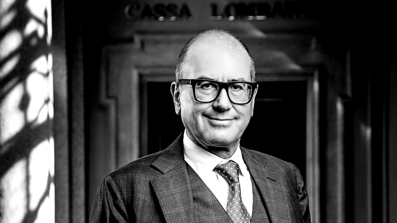 Una persona in giacca e cravatta, con gli occhiali, sta in piedi con sicurezza davanti a una porta decorata con "Cassa Lombarda" sottilmente visibile sullo sfondo. L'immagine è in bianco e nero, catturando un'eleganza senza tempo che ricorda lo stile distintivo di Vistalli.