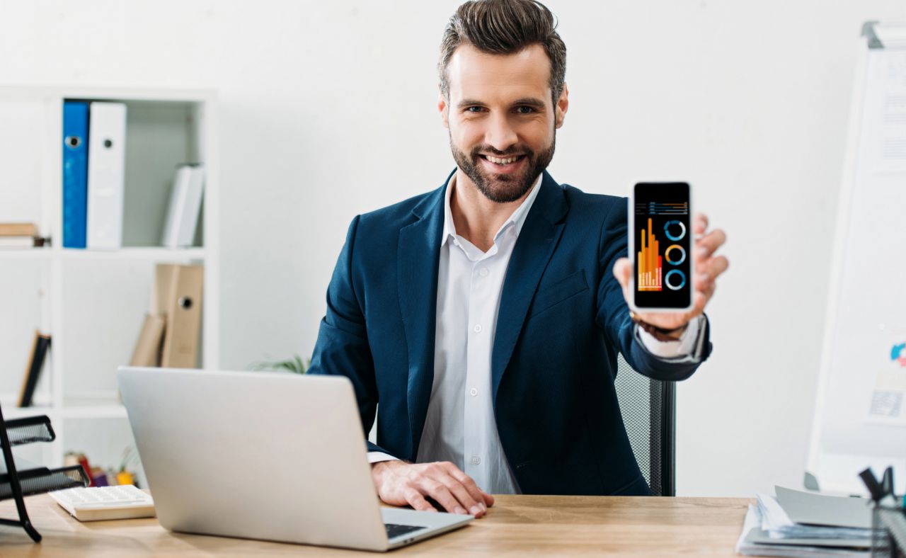 Un uomo in giacca e cravatta è seduto a una scrivania con un computer portatile, sorride e tiene in mano uno smartphone su cui sono visualizzati grafici e tabelle. Sullo sfondo si vedono librerie e articoli da ufficio.