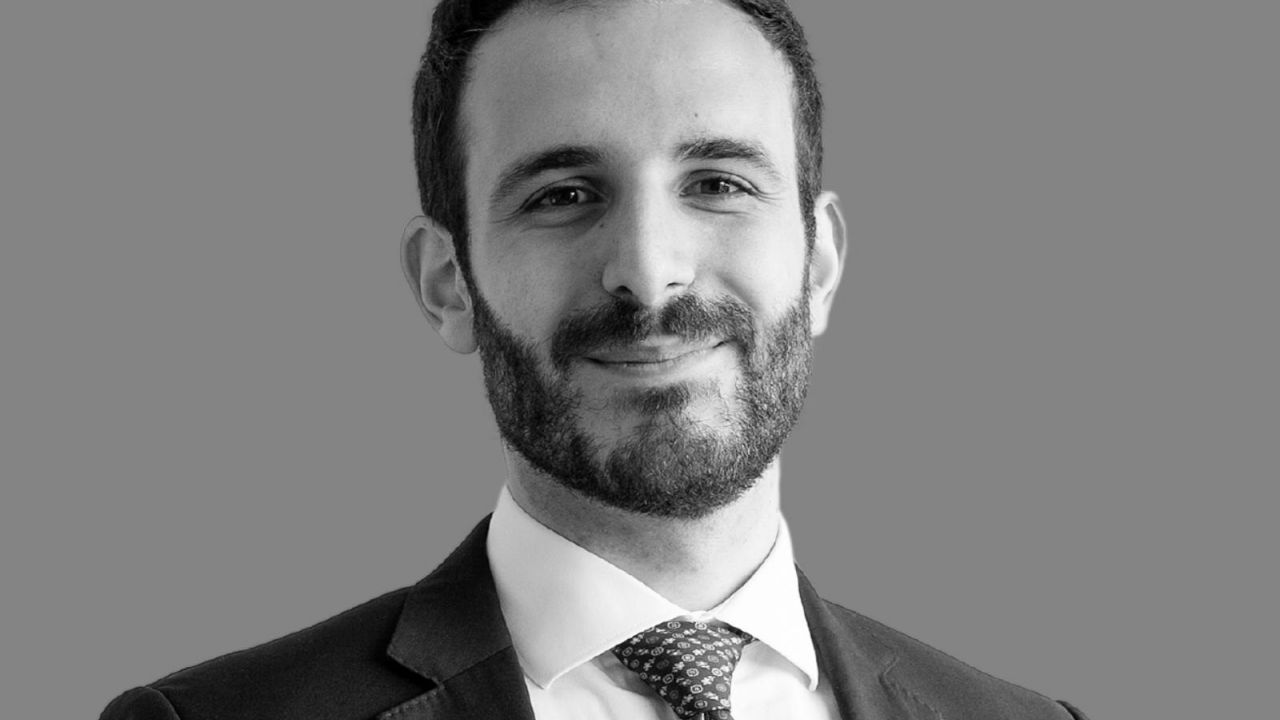 Foto in bianco e nero di un uomo con la barba, in giacca e cravatta, sorridente su uno sfondo grigio.