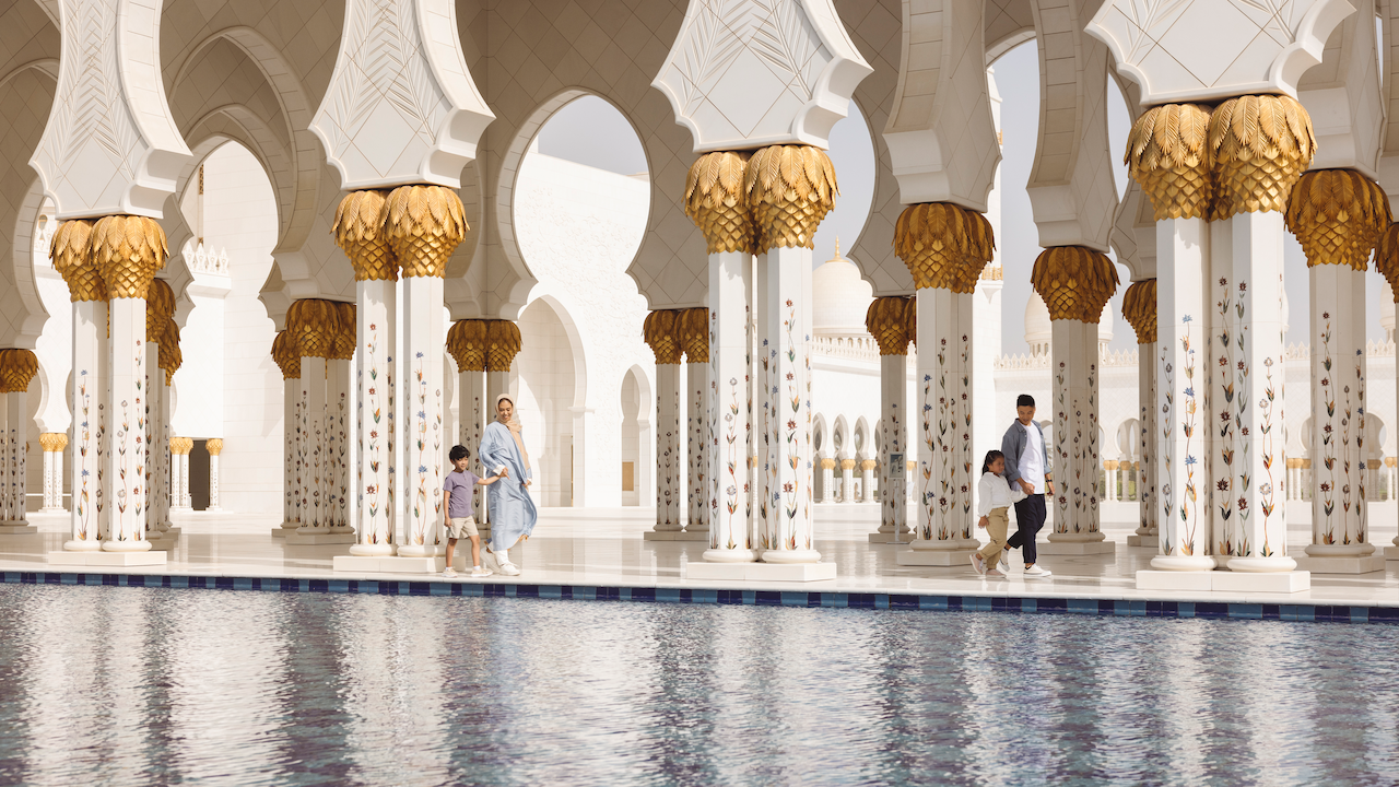 La Gran moschea dello Sceicco Zayed. Una famiglia con bambini piccoli cammina lungo un corridoio di marmo lucido decorato con intricate colonne floreali, vicino a una piscina riflettente in una grande moschea. L'architettura presenta cupole bianche e accenti dorati, con archi che offrono scorci del cielo.