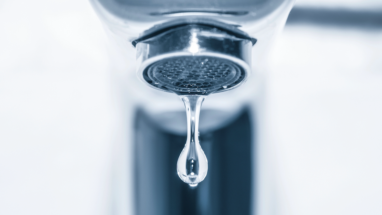 Investire nell'acqua. Primo piano di un rubinetto con una singola goccia d'acqua che cade dal beccuccio. Lo sfondo è leggermente sfocato, evidenziando la goccia e la texture metallica del rubinetto.