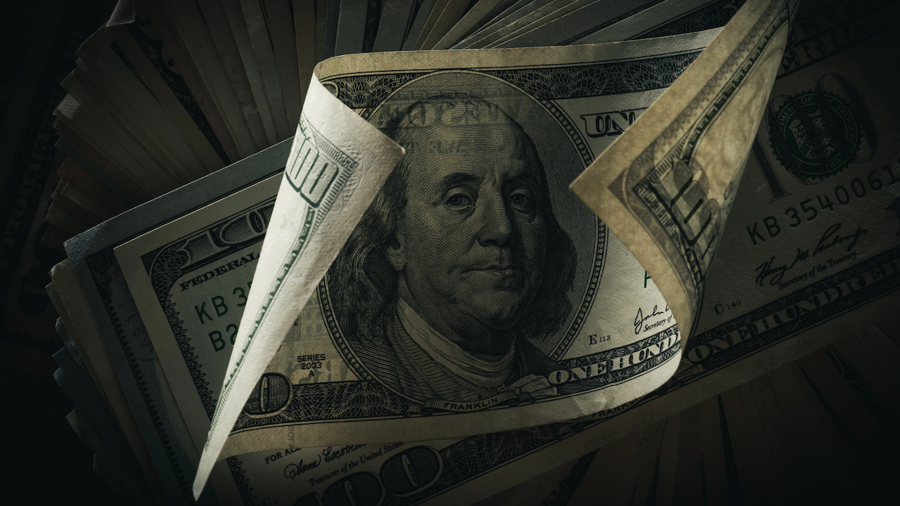 Un primo piano di una pila di banconote da cento dollari statunitensi, con in primo piano il ritratto di Benjamin Franklin. Le banconote sono leggermente disposte a ventaglio, con una banconota che si arriccia all'angolo, creando ombre e mettendo in risalto i dettagli intricati.