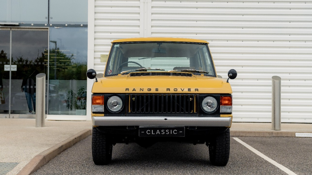 Una Range Rover gialla classica è parcheggiata di fronte a un edificio moderno con pareti bianche e grandi finestre in vetro. Il veicolo d'epoca ha una targa "Classic" sulla parte anteriore. Lo sfondo mostra un sentiero e parte della strada.