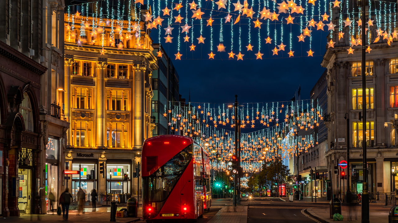 Una scena di strada vivace di notte a Londra, con luci festive a forma di stella dall'altra parte della strada. È visibile un autobus rosso a due piani e gli edifici sono illuminati su entrambi i lati, incluso un negozio con il logo Microsoft.
