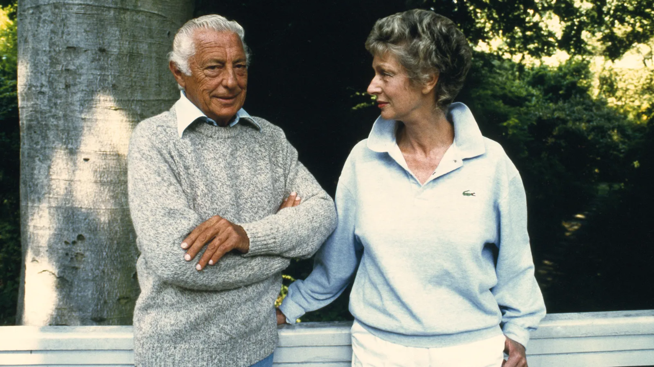 Un uomo e una donna anziani sono in piedi all'aperto, sorridenti l'uno all'altra. L'uomo indossa un maglione grigio e la donna indossa un pullover azzurro. Sono in una zona verdeggiante, appoggiati a una ringhiera bianca.