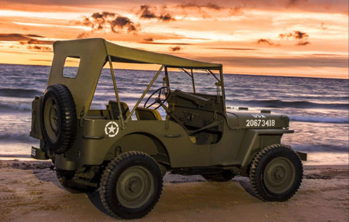 Una jeep militare d'epoca è parcheggiata su una spiaggia sabbiosa al tramonto. Il veicolo, contrassegnato con il numero 20673418 e una stella bianca, è rivolto verso l'oceano mentre le onde lambiscono dolcemente la riva sotto un cielo arancione e rosa vibrante.