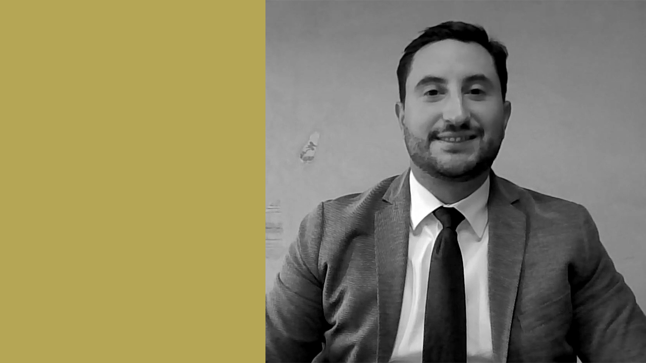 Una foto in bianco e nero di un uomo in giacca e cravatta, sorridente, su uno sfondo semplice. Il terzo sinistro dell'immagine è di un solido color senape, mentre il resto mostra l'uomo dal petto in su.