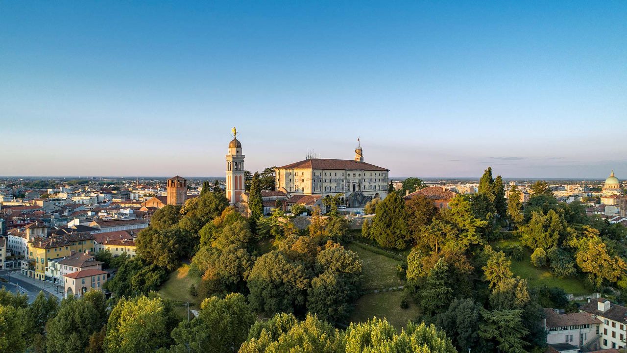Una vista panoramica di una città storica con un importante edificio caratterizzato da una torre dell'orologio e una cupola, circondato da una vegetazione lussureggiante. Lo skyline è ampio, con un cielo azzurro terso sopra la testa.