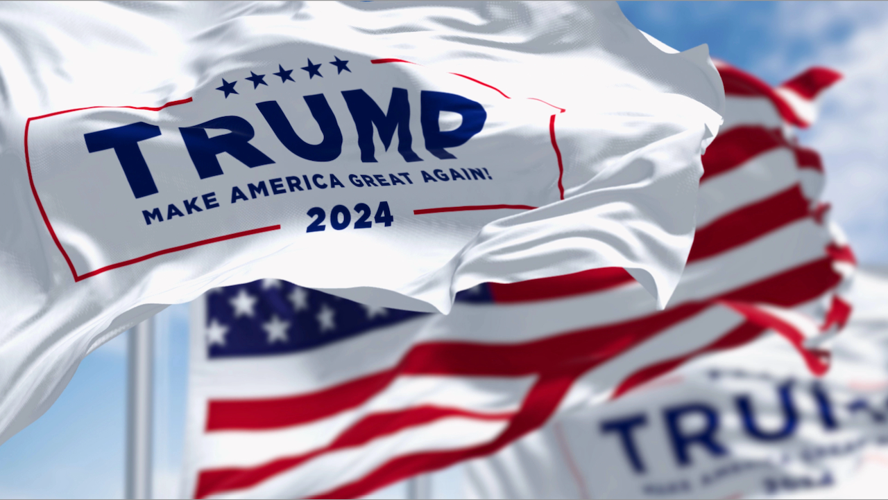 Bandiere bianche con la scritta "TRUMP Make America Great Again! 2024" sventolano al vento, contro un cielo azzurro. Sullo sfondo sono visibili le bandiere americane.