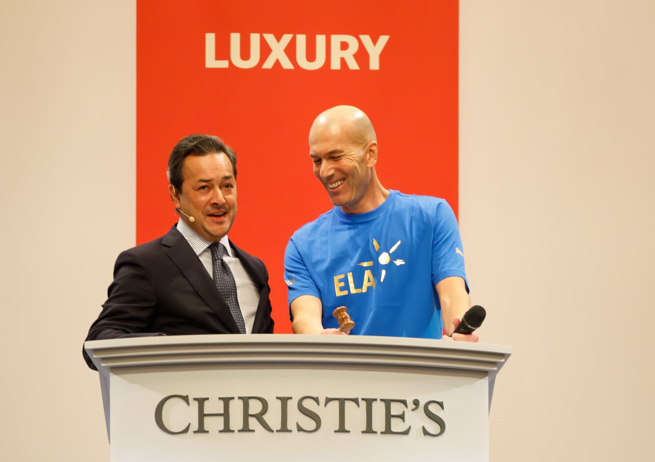 Zidane con il capo degli orologi di Christie's Europa e Usa è in piedi sul podio di un'asta di Christie's. Uno è vestito in completo e l'altro indossa una maglietta blu. Sono entrambi sorridenti, con uno sfondo rosso con la scritta "LUXURY" in lettere bianche. L'uomo con la maglietta tiene in mano un martelletto.