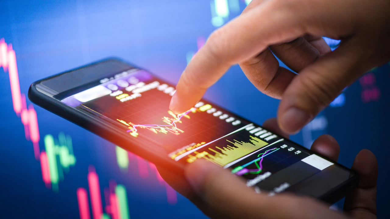Una persona che interagisce con uno smartphone mostrando i dati di borsa di Fideuram Direct, completi di grafici e diagrammi, su uno sfondo sfocato di informazioni finanziarie su uno schermo ampio. L'immagine fonde perfettamente temi di finanza e tecnologia.