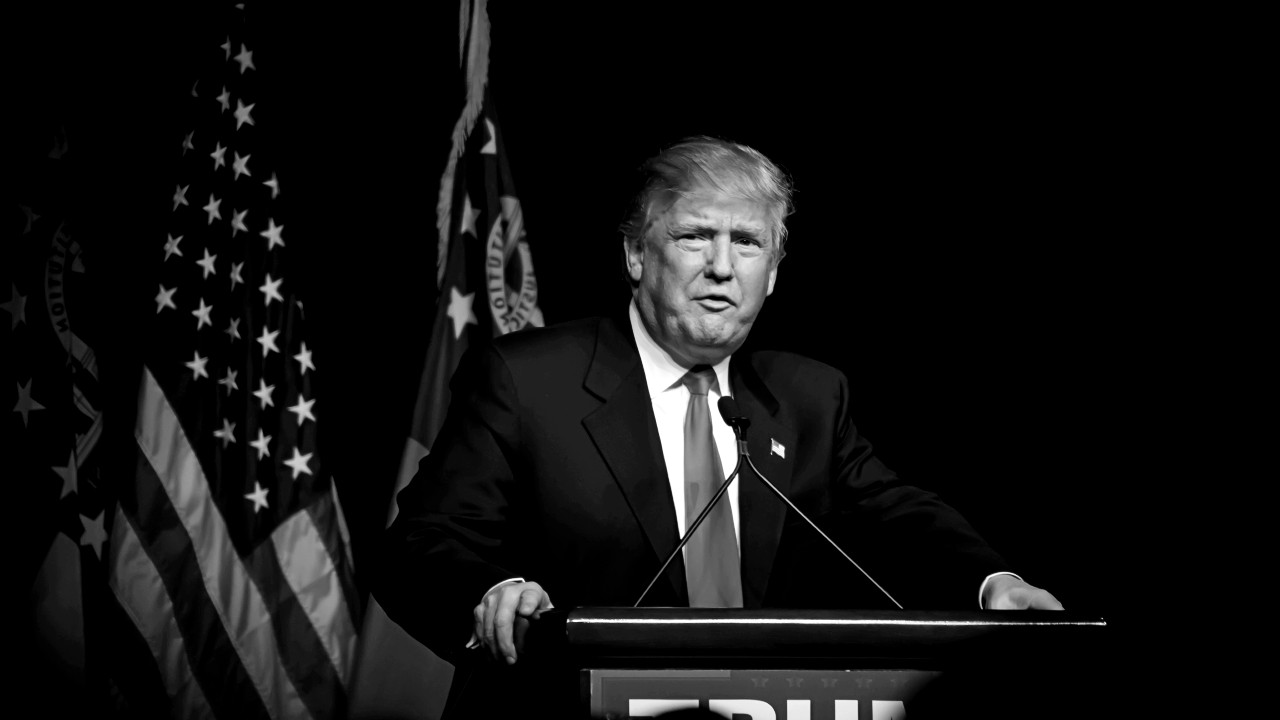 Immagine in bianco e nero di Donald Trump in giacca e cravatta, in piedi su un podio, mentre si rivolge al pubblico. Sullo sfondo una bandiera americana. In vista delle elezioni, quale potrebbe essere l'impatto sui Btp e sugli altri titoli di stato?
