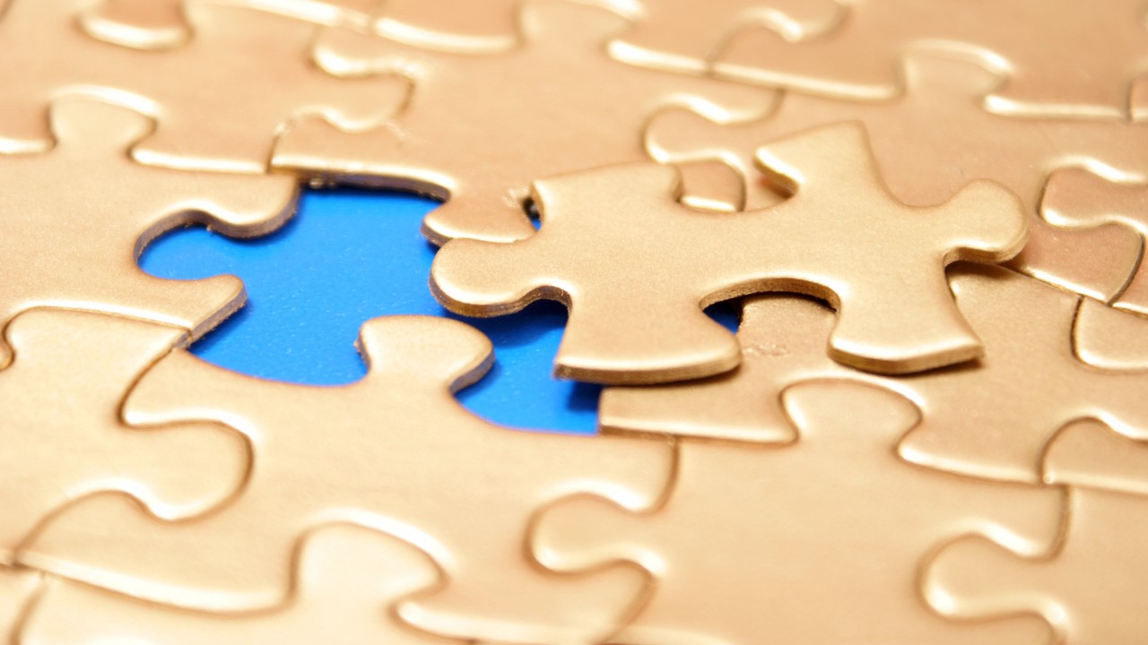 Primo piano di un puzzle d'oro parzialmente completato con un pezzo mancante, che rivela uno sfondo blu brillante sottostante. Un singolo pezzo aleggia sopra lo spazio vuoto, pronto per il conferimento all’interno del puzzle.
