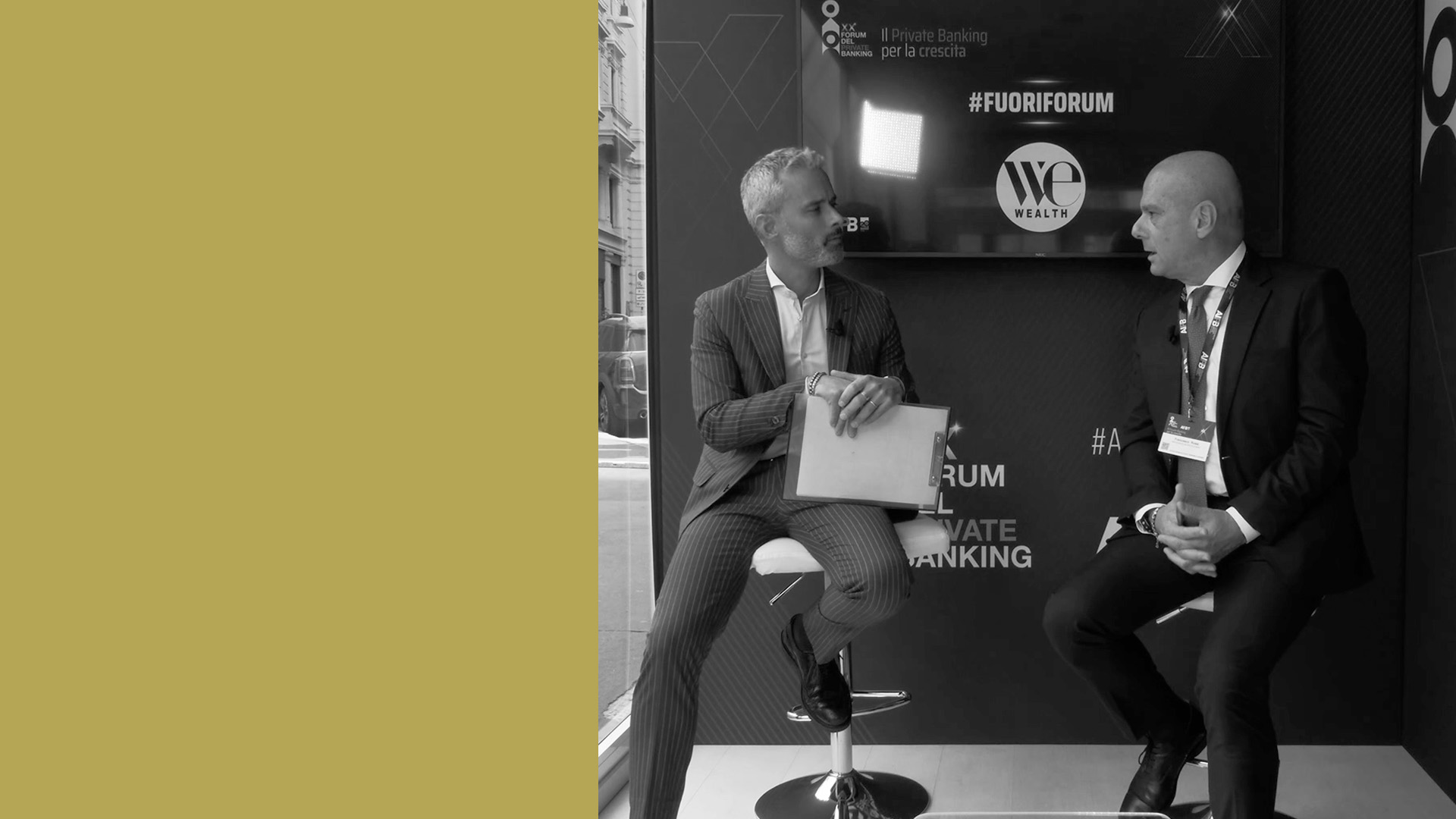 Due uomini in abiti da lavoro sono seduti su degli sgabelli e stanno conversando. Sono di fronte a uno schermo che mostra i loghi "Fuoriforum" e "We Wealth". L'ambientazione sembra essere un evento professionale o un'intervista. Forum AIPB 2024