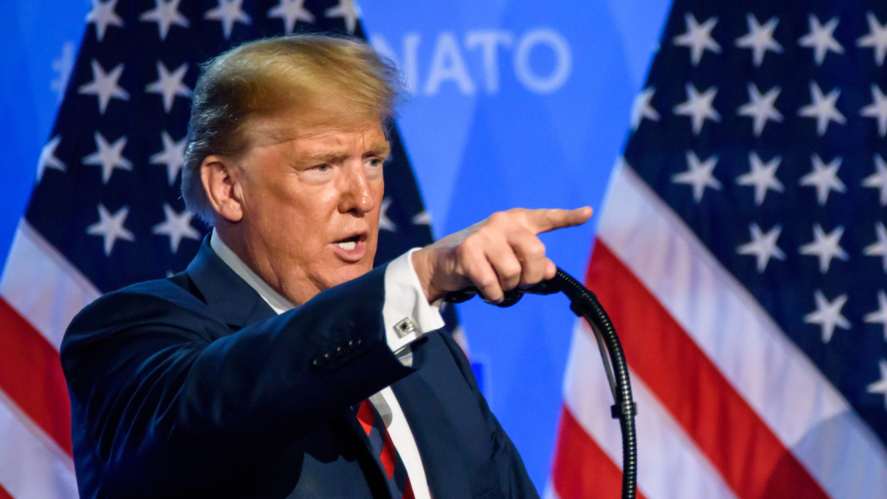 Un uomo in giacca e cravatta, che ricorda l'atteggiamento sicuro di sé di Trump, gesticola mentre parla da un podio con due bandiere americane sullo sfondo e la parola "NATO" parzialmente visibile.