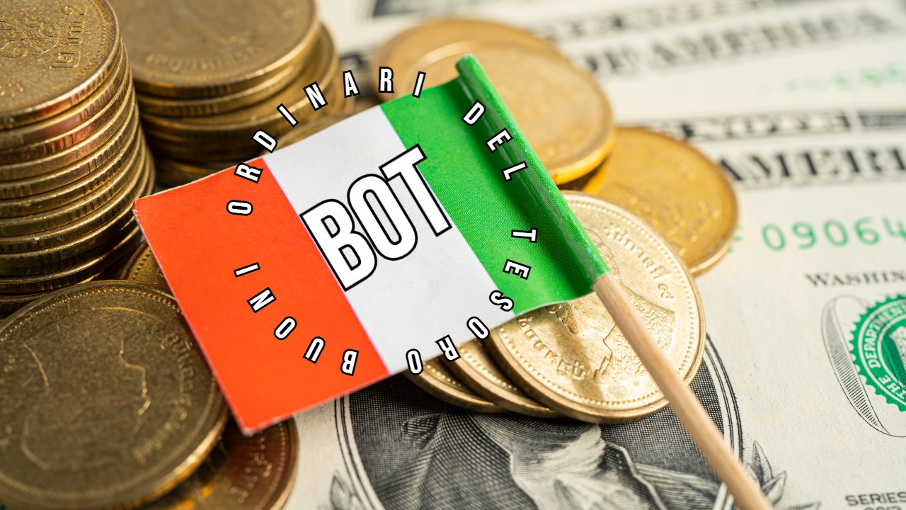 Una piccola bandiera con i colori italiani e la scritta "BOT" circondata dal testo "Buoni Ordinari del Tesoro" è posta su una pila di monete e banconote da un dollaro statunitense.