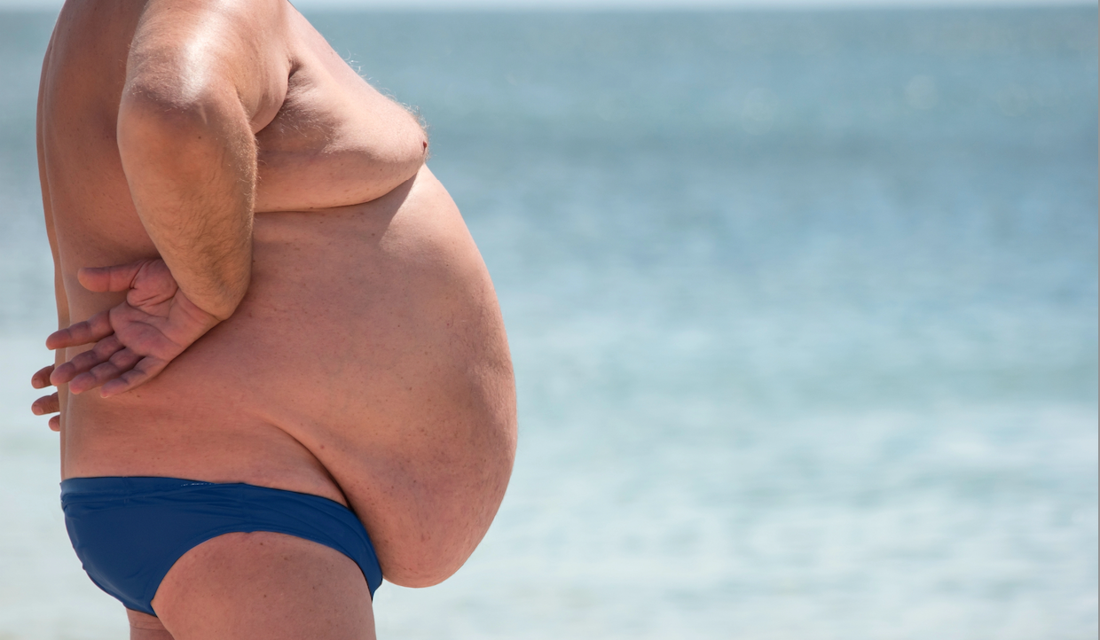 Una persona in sovrappeso che indossa un costume da bagno blu è in piedi sulla spiaggia con la schiena girata, una mano sul fianco. L'attenzione è rivolta al suo torso contro uno sfondo sfocato dell'oceano.