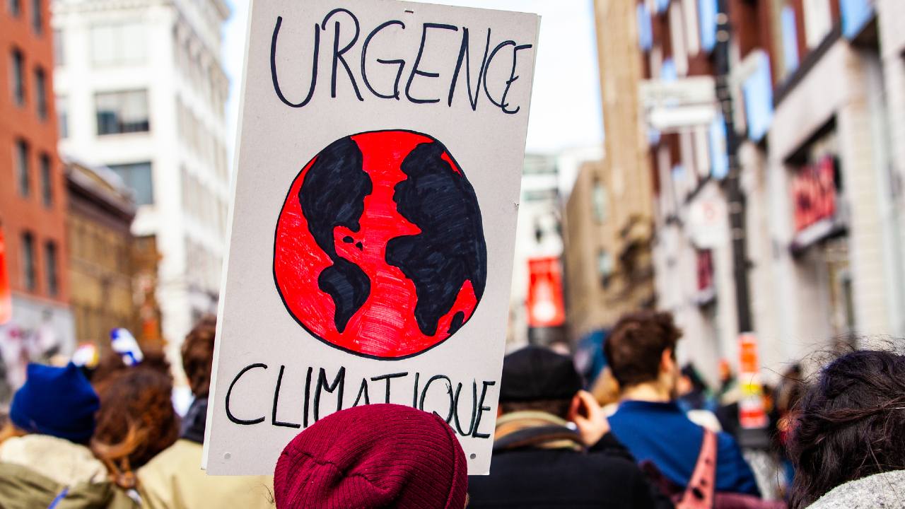 I manifestanti in una marcia cittadina con un cartello che recita "Urgence Climatique", raffigurante un'illustrazione della Terra rossa e nera. L'urgenza della crisi climatica ha portato molti a mettere in discussione l'assegnazione di fondi agli sforzi ambientali, mentre edifici sfocati e folle formano lo sfondo.