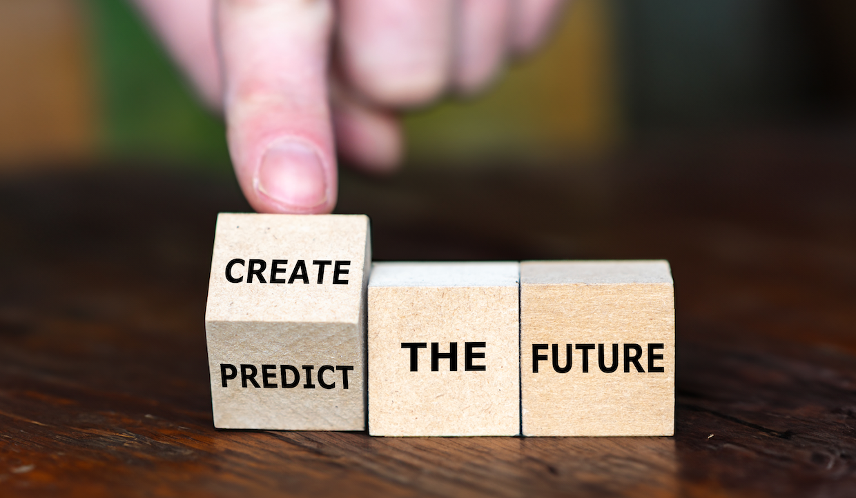 Una mano sta capovolgendo un cubo di legno con le parole "PREDICT" e "CREATE" sui lati. Altri due cubi riportano la scritta "THE FUTURE". La trasformazione suggerisce un cambiamento dal predire il futuro al crearlo. Lo sfondo è una superficie di legno.