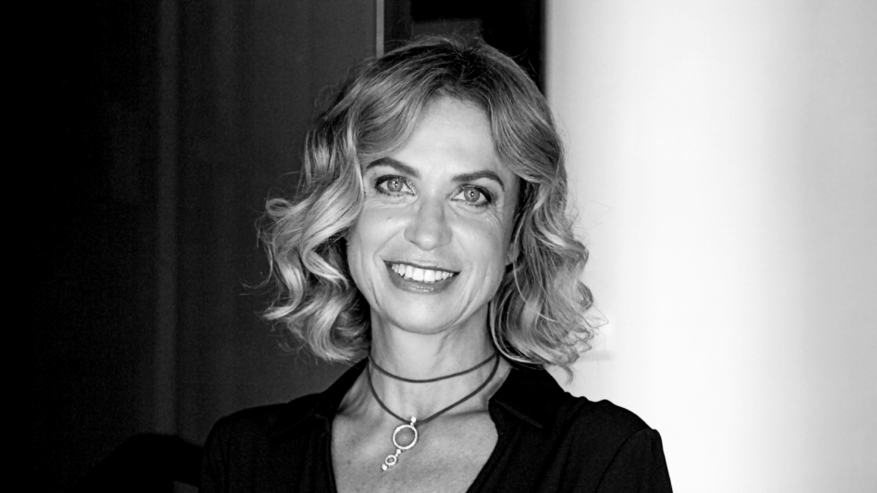 Ritratto in bianco e nero di una donna con i capelli mossi, che sorride dolcemente alla telecamera. Vestita con un top scuro, indossa una collana con un pendente circolare che accenna sottilmente al suo matrimonio. Lo sfondo è delicatamente sfocato.