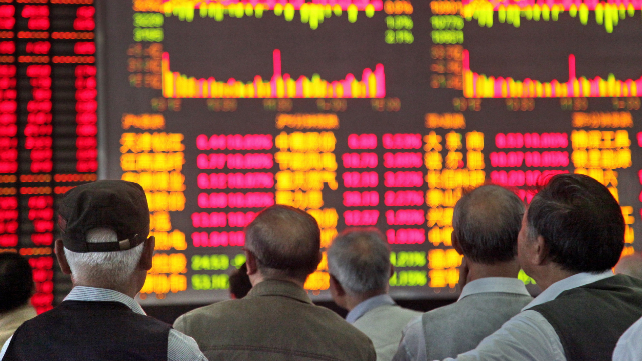 Un gruppo di persone sta di fronte a un grande display digitale, probabilmente della Borsa di Shanghai, che mostra i dati del mercato azionario con numeri e grafici rossi e gialli. Lo schermo presenta vividamente informazioni e tendenze finanziarie.