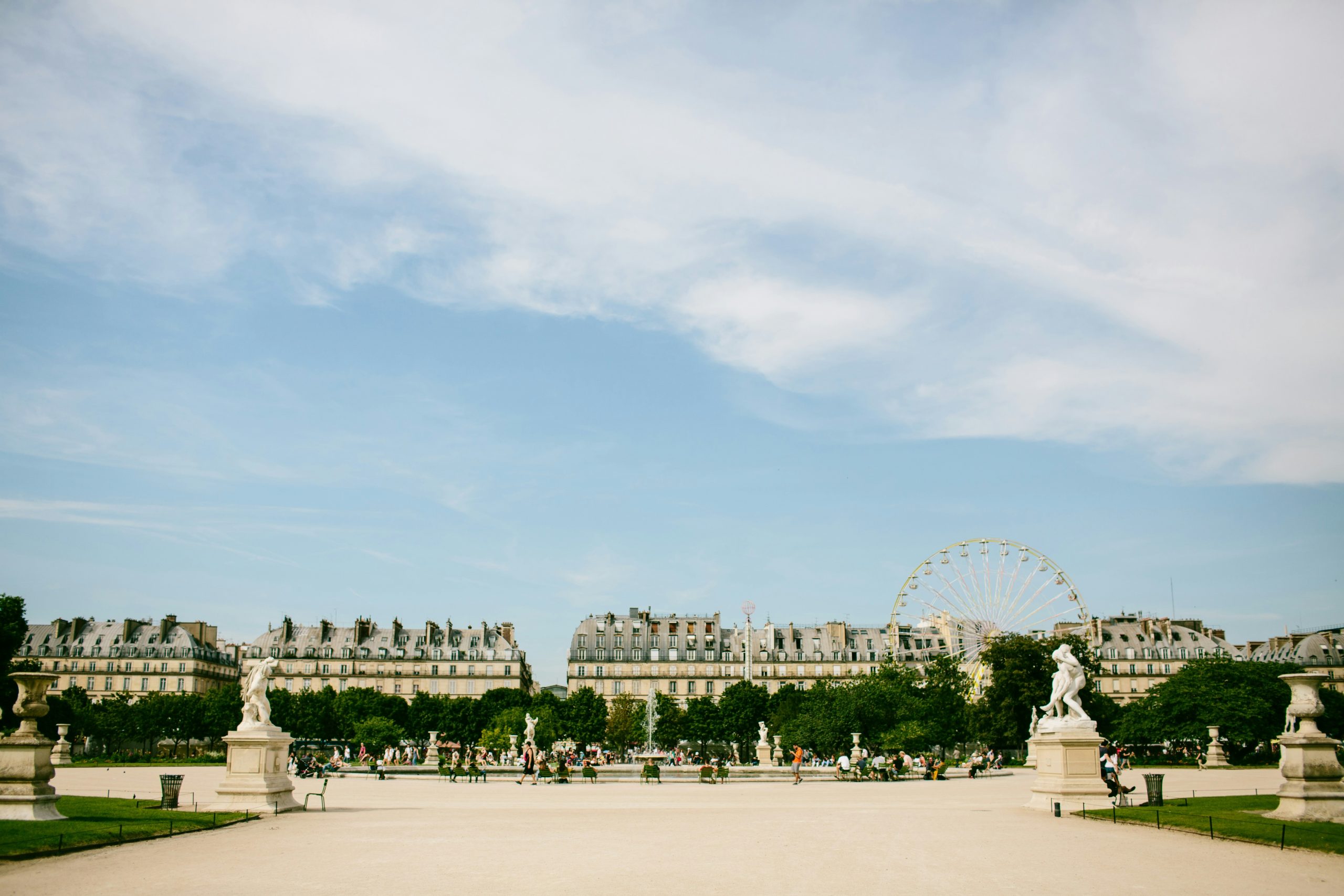 Una giornata di sole al Giardino delle Tuileries a Parigi, con persone che camminano lungo i sentieri. Statue ornate fiancheggiano il sentiero e una ruota panoramica si erge alta sullo sfondo contro un cielo azzurro terso. Edifici storici costeggiano l'orizzonte.