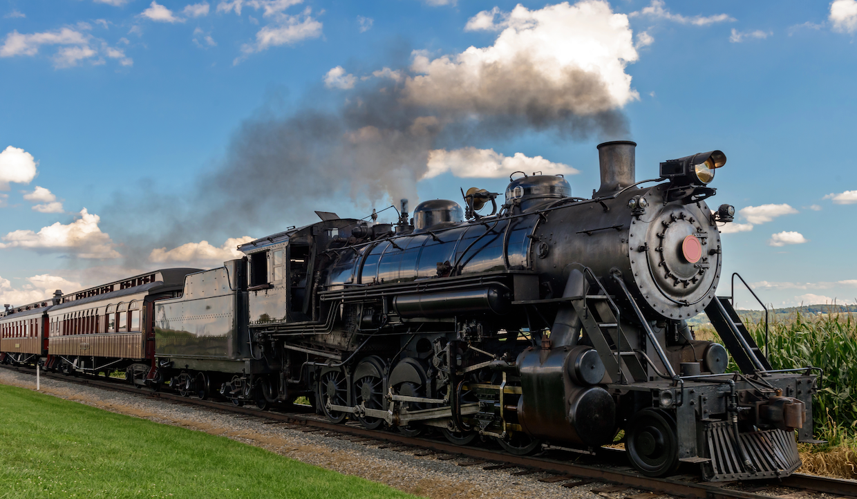 Una locomotiva a vapore d'epoca con un esterno nero lucido traina una fila di carrozze passeggeri classiche. Un denso fumo nero si solleva dalla ciminiera contro uno sfondo di cielo azzurro e nuvole sparse. Erba verde e campi di mais costeggiano i binari.