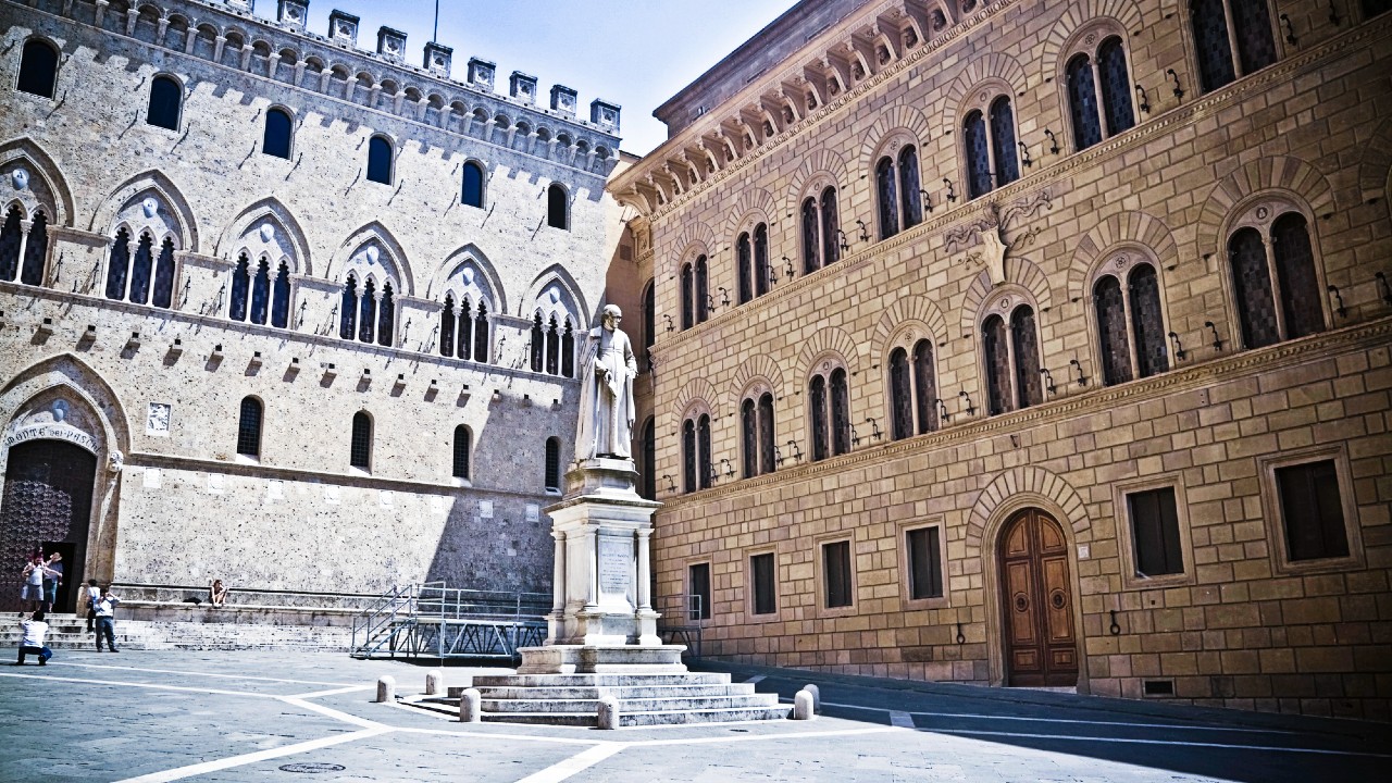 Una piazza storica con una statua di una figura in piedi su un piedistallo di fronte a due grandi edifici in pietra ornati con finestre ad arco. Gli edifici mostrano un'architettura classica con dettagli intricati. Il cielo è limpido e soleggiato.