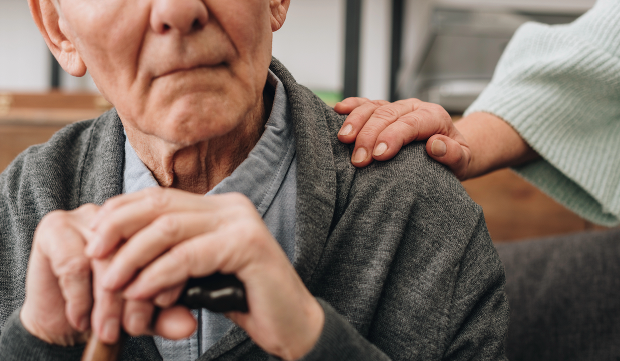 Una persona anziana appoggia le mani su un bastone, indossando un maglione grigio. La mano di un'altra persona è appoggiata delicatamente sulla sua spalla, offrendole supporto e conforto. La scena trasmette cura e compagnia.