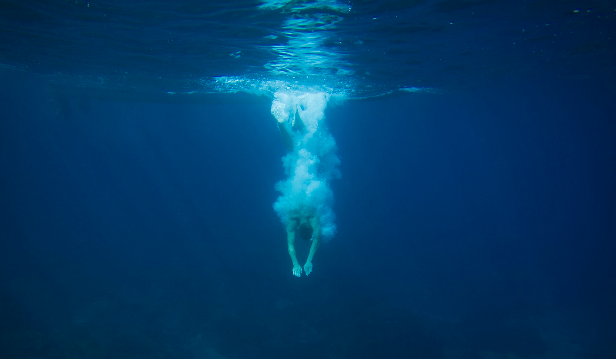Una persona si tuffa in acque blu intenso, creando schizzi e una scia di bolle mentre scende dalla superficie. La luce del sole filtra attraverso l'acqua, illuminando la scena.