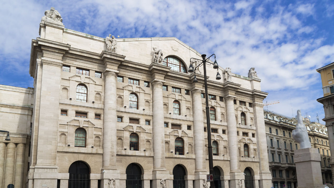 L'immagine cattura un grandioso edificio neoclassico, che ricorda l'eleganza architettonica di Piazza Affari, con sculture ornate sul tetto, grandi colonne e finestre ad arco. È una giornata luminosa, parzialmente nuvolosa, e l'edificio è circondato da altre strutture.