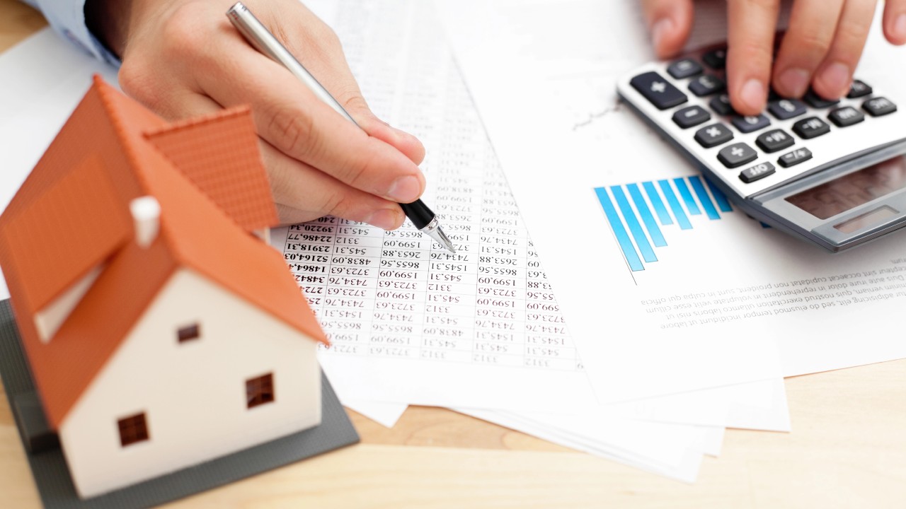 Una persona usa una calcolatrice e scrive su documenti con numeri e grafici a barre. Un piccolo modello di casa è appoggiato sul tavolo, suggerendo calcoli finanziari incentrati sull'immobiliare relativi al settore immobiliare.