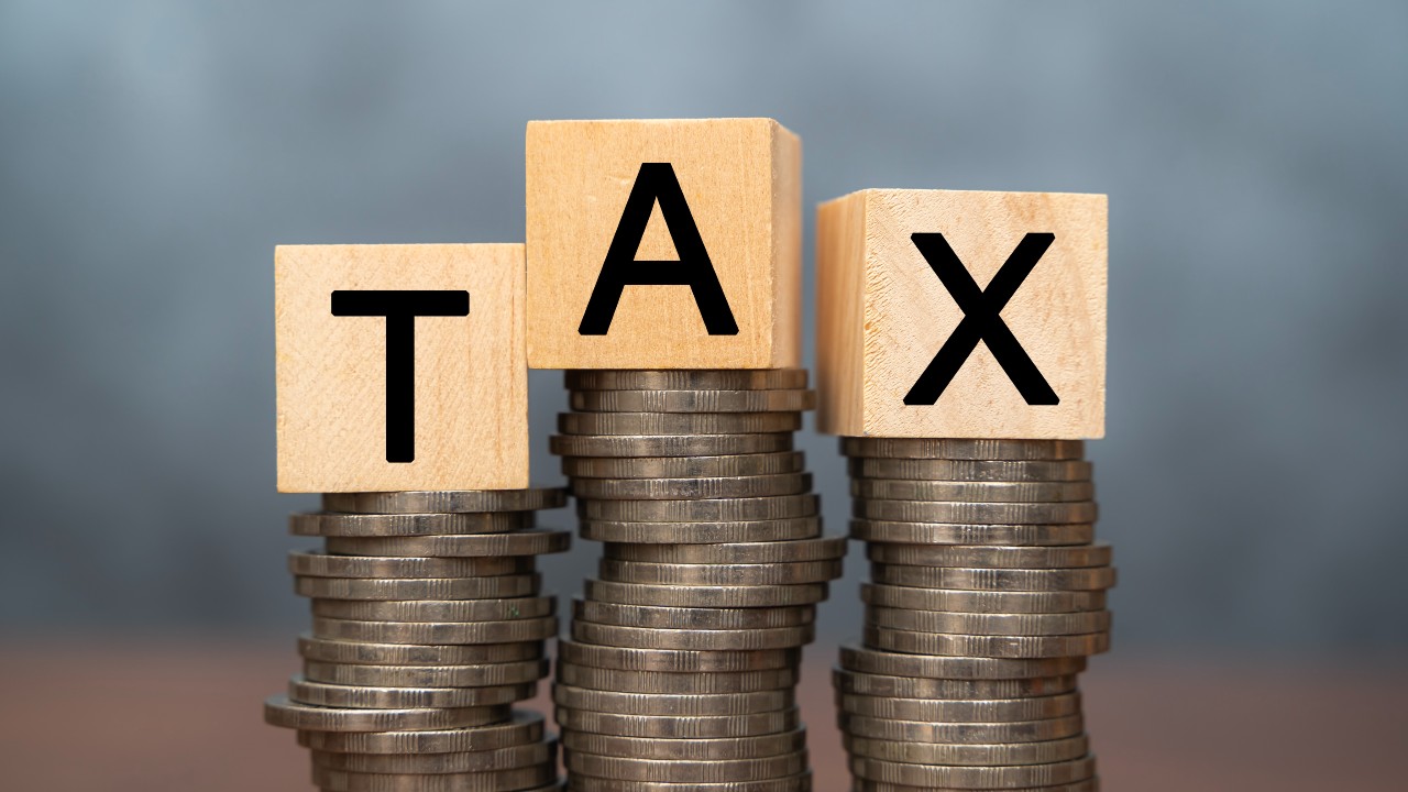 Blocchi di legno con la scritta "TAX" sono posizionati sopra pile di monete su uno sfondo blu-grigio sfocato, a simboleggiare l'intersezione tra ricchezza e obblighi finanziari.