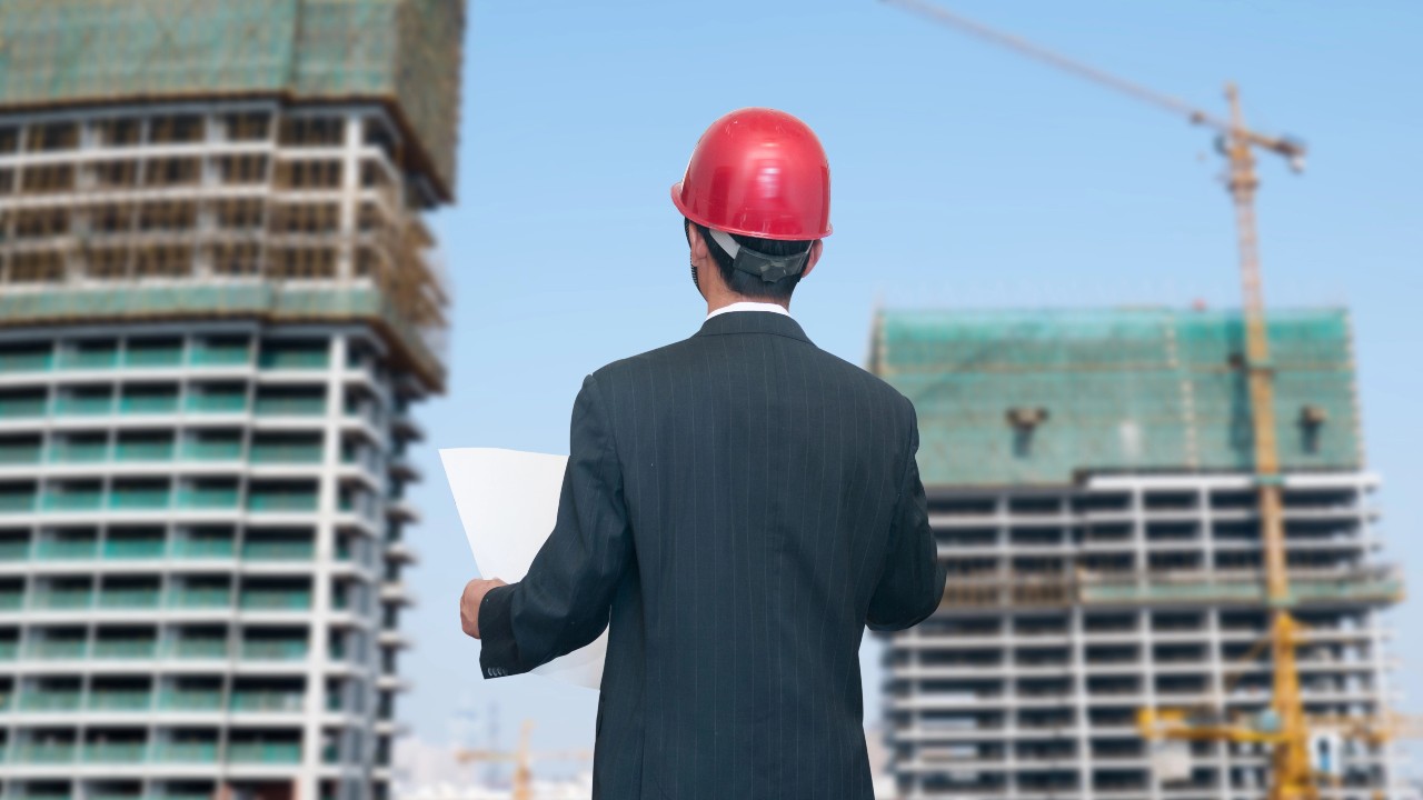 Un uomo in giacca e cravatta e casco rosso tiene in mano delle planimetrie, in piedi di fronte a due grattacieli in costruzione, con le gru che si stagliano contro un cielo azzurro e terso. L’immagine vuole riportare al mondo immobiliare e al trattamento fiscale da applicare alle somme risarcitorie nei contratti di appalto