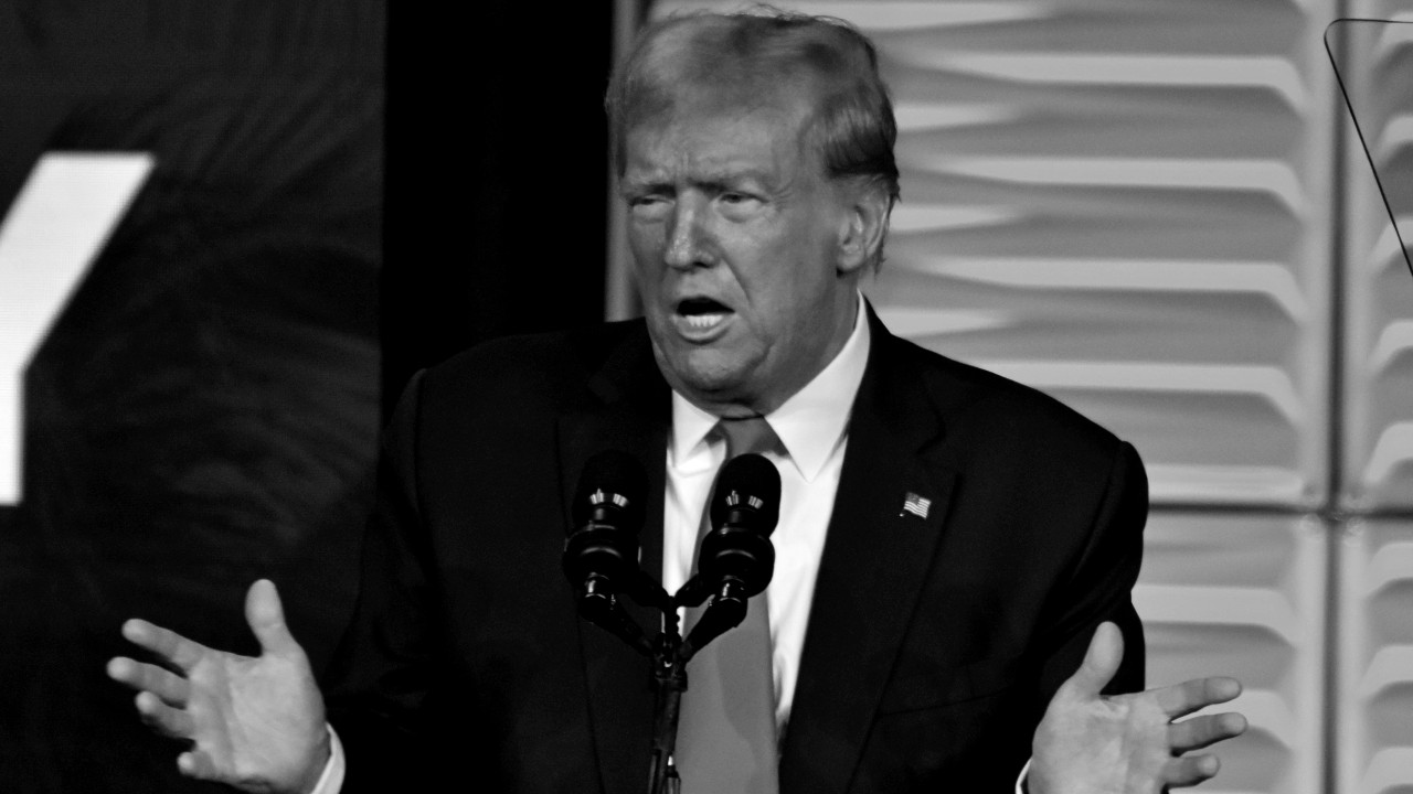 Il presidente degli Usa, Donald Trump, in giacca e cravatta, gesticola con le mani aperte mentre parla in più microfoni. I pannelli testurizzati sullo sfondo bianco e nero aggiungono profondità alla scena.