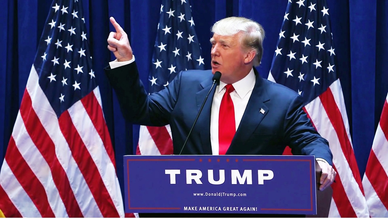 Donald Trump, in giacca e cravatta rossa parla da un podio su cui è inciso il suo nome. Fa un gesto verso l'alto, in segno di vittoria, con la mano destra, mentre sullo sfondo sventolano orgogliosamente diverse bandiere americane.
