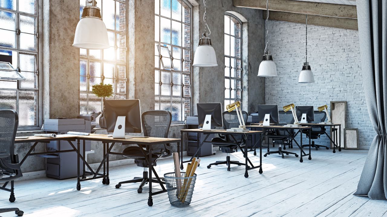 Uno spazio moderno per uffici con ampie finestre che lasciano entrare la luce naturale, con diverse postazioni di lavoro dotate di computer. La stanza fonde design industriale e minimalista, vantando pavimenti in legno, pareti in mattoni a vista e lampade a sospensione. L'immagine vie usata per parlare di uffici flessibili e spazi condivisi e di come cambia il lavoro in azienda.