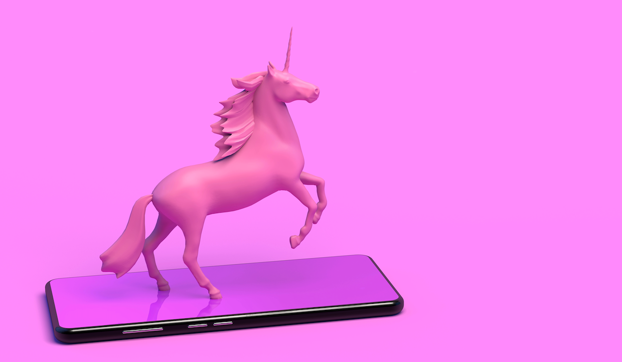Un unicorno rosa che emerge da uno smartphone, su uno sfondo rosa acceso. Il telefono è appoggiato in piano e l'unicorno si sta sollevando sulle zampe posteriori, creando una scena fantasy moderna e stravagante.