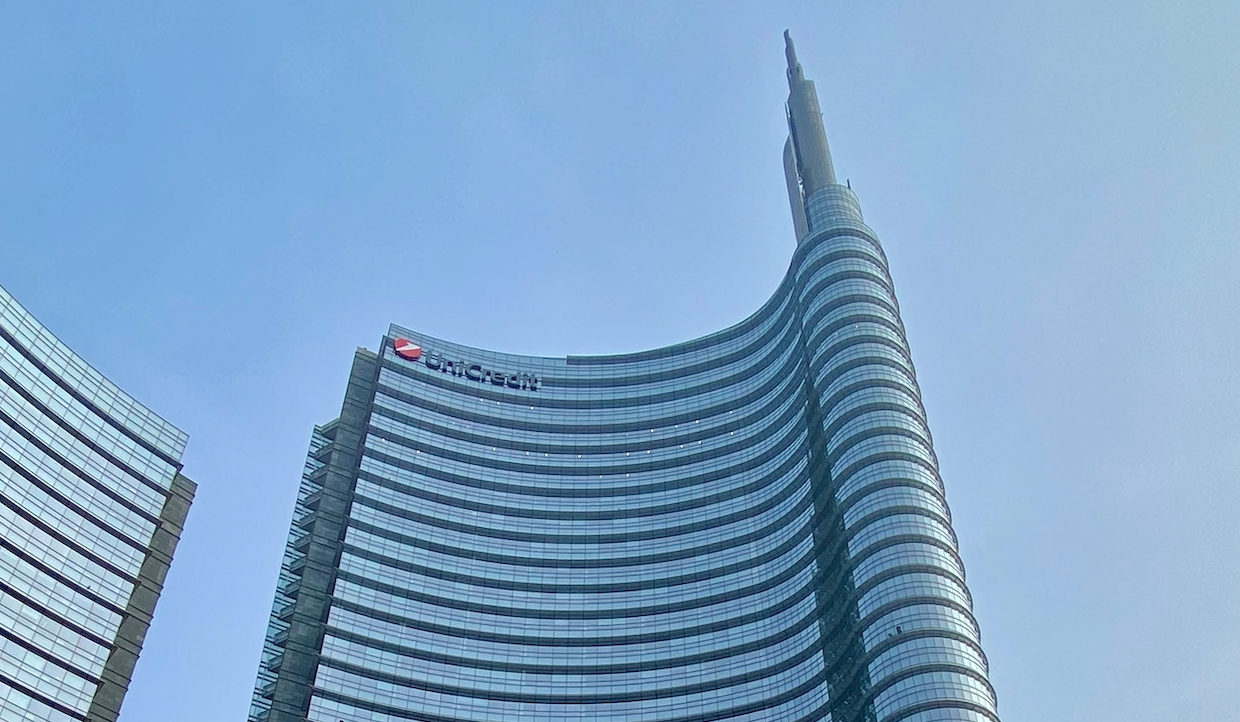 Un grattacielo moderno e curvo con una guglia, caratterizzato da finestre in vetro riflettente. La facciata dell'edificio mostra il logo UniCredit. Il cielo è limpido e azzurro, il che enfatizza l'altezza dell'edificio e il suo design architettonico elegante.