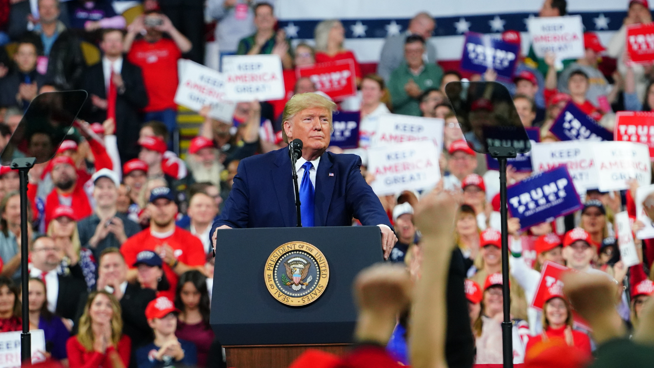 Un uomo in giacca e cravatta è in piedi su un podio con il sigillo presidenziale, mentre si rivolge a una folla entusiasta. Tra cori e applausi, i membri del pubblico tengono cartelli con la scritta "Keep America Great" e "Trump", indossando con orgoglio cappelli e camicie rosse mentre si radunano dietro Donald Trump, alimentando un'atmosfera energica.