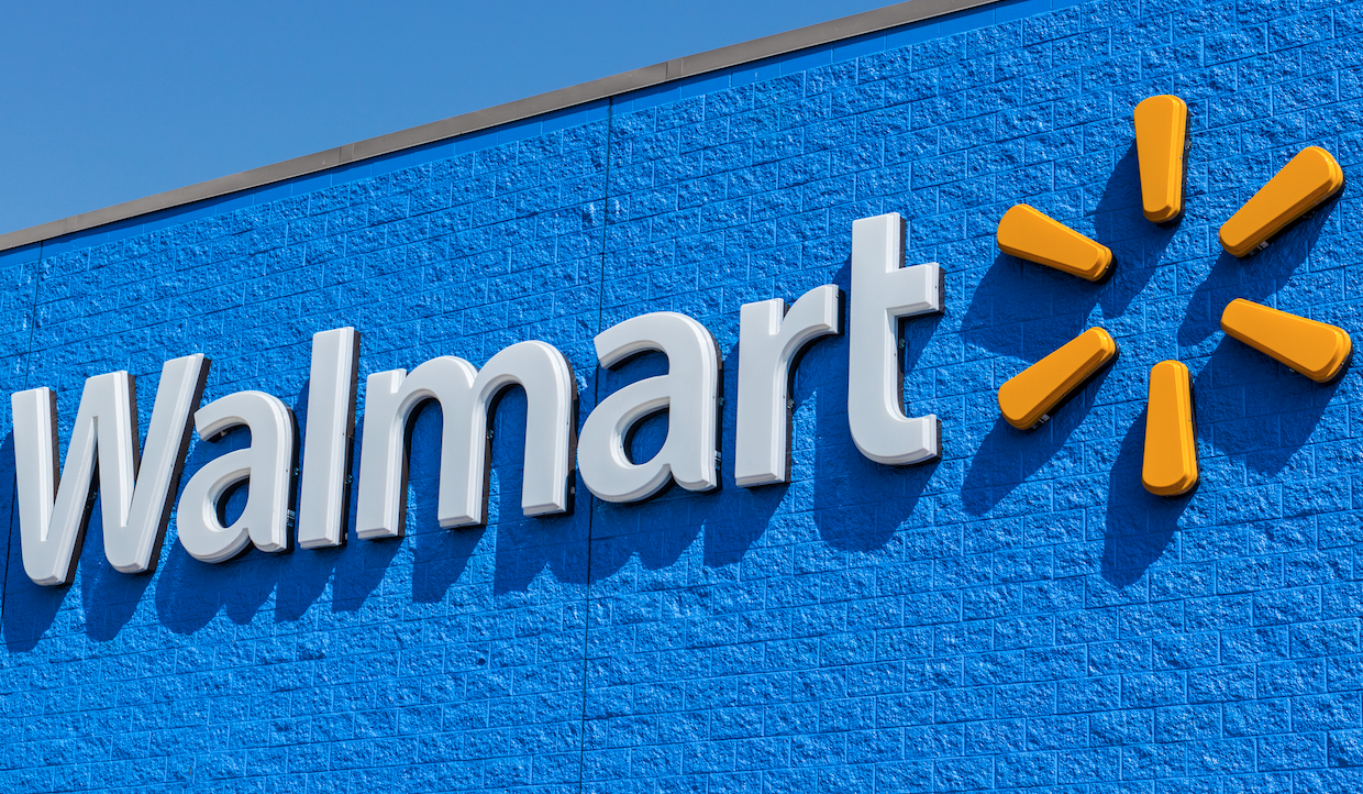L'immagine presenta l'iconico logo Walmart in bella mostra su un muro di mattoni blu brillante. La parola "Walmart" appare in lettere bianche nitide, accompagnata da un simbolo a raggiera realizzato con forme gialle sul lato destro, che incarna l'essenza vibrante del marchio.