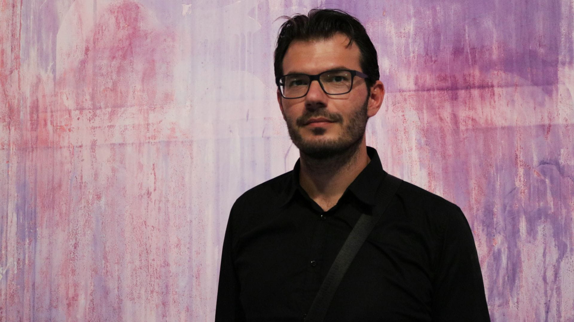 Un uomo con gli occhiali e una camicia nera è in piedi davanti a uno sfondo astratto viola e rosa.