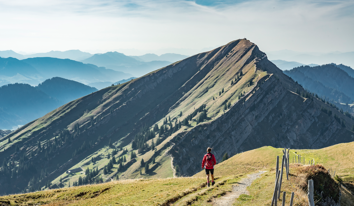 Una persona che indossa una giacca rossa cammina lungo uno stretto sentiero che conduce verso una ripida cresta montuosa erbosa. Il cielo è limpido e sullo sfondo si vedono strati di montagne lontane e nebbiose sotto un cielo azzurro tenue.