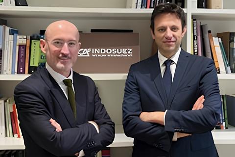 Due uomini in giacca e cravatta sono in piedi, con le braccia incrociate, davanti a una libreria con vari libri e un cartello con la scritta "INDOSUEZ". Entrambi sorridono leggermente e sono in posa in un ambiente professionale.